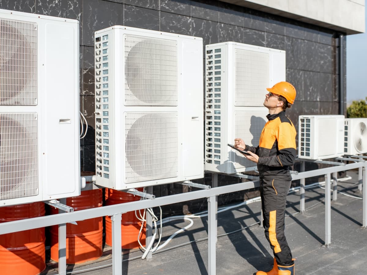 Technician in safety gear examining multiple outdoor air conditioning condenser units on a building rooftop