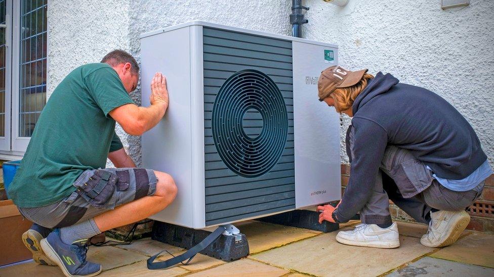 Two technicians positioning a large outdoor heat pump unit beside a residential wall