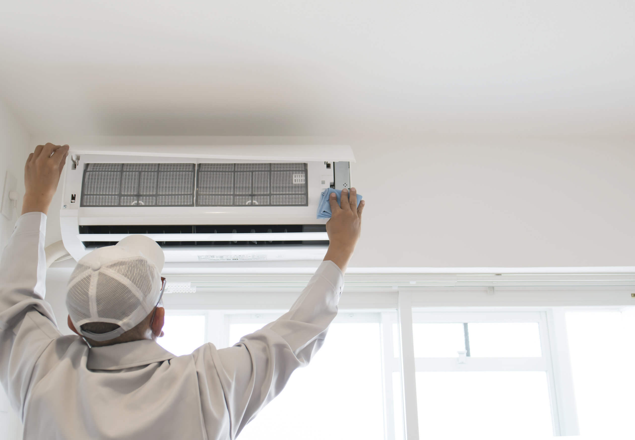 Technician wiping the filter area of a wall-mounted air conditioning unit inside a bright residential room
