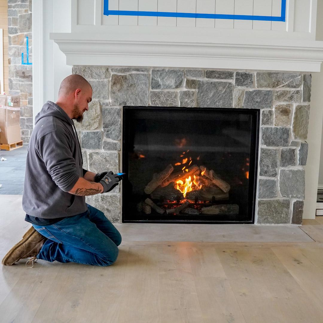 Technician kneeling to inspect a lit gas fireplace with a stone surround in a modern living room